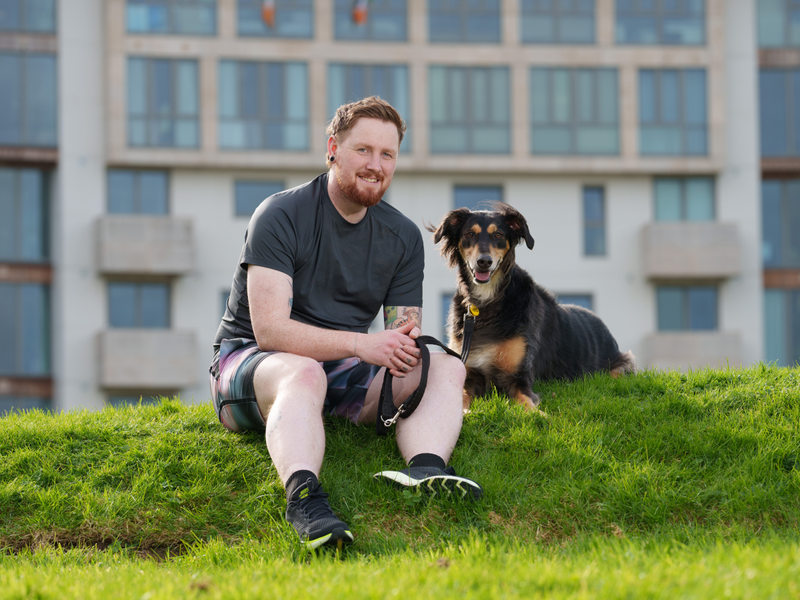 Man in running wear sits on grassy hill with a black and tan saluki type dog